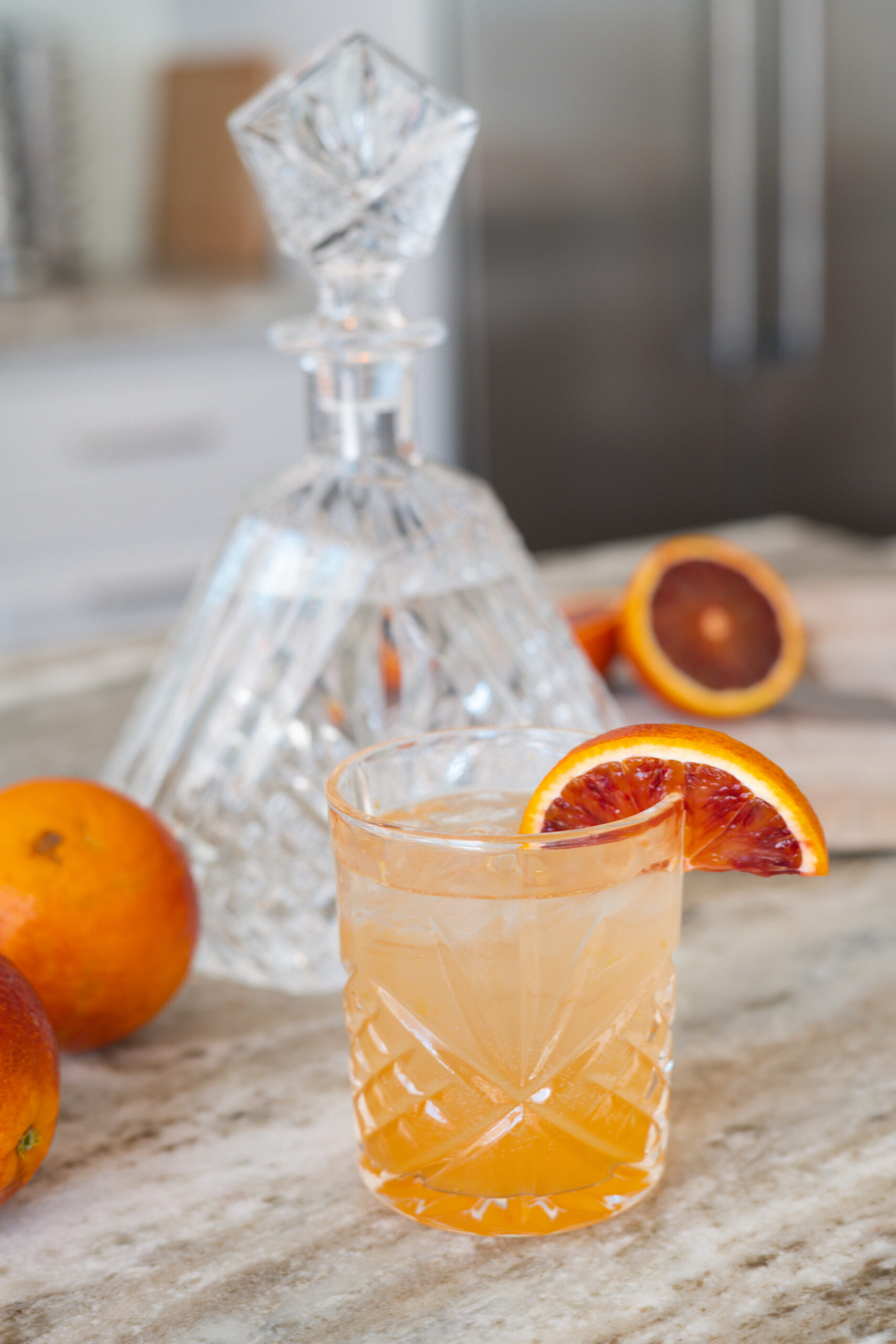 Short cocktail glass with an orange drink and a blood orange slice. There are blood orange slices in the background with a crystal decanter.