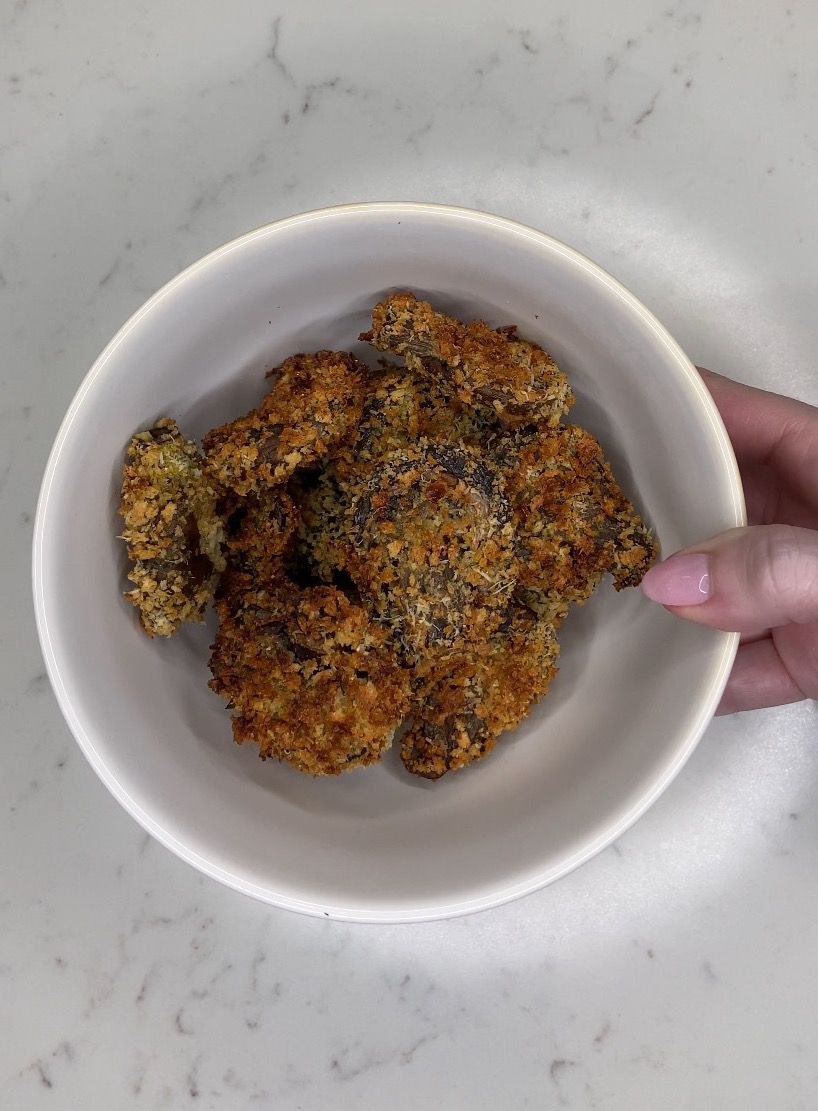 Mushrooms coated in breadcrumbs and Parmesan cheese that have just been air fried and are in a white bowl and a hand with pink nails is holding it.